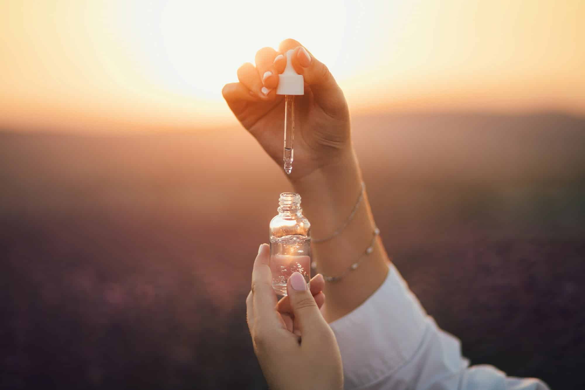 Close up female hands holding essential oil in a bottle with pipette in lavender field at sunset.